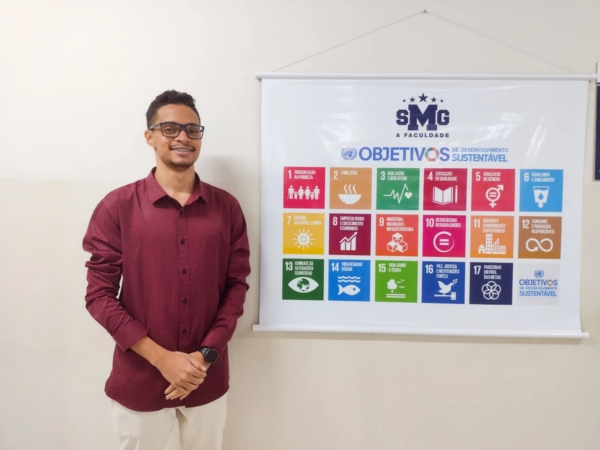 Thiago Felipe Moreira de Almeida, aluno de Engenharia da UNISMG, em foto ao lado do banner com os 17 ODS. O acadêmico desenvolveu uma telha ecológica, inspirado pelo ODS 7. Foto: Sthefany Macedo/UNISMG.